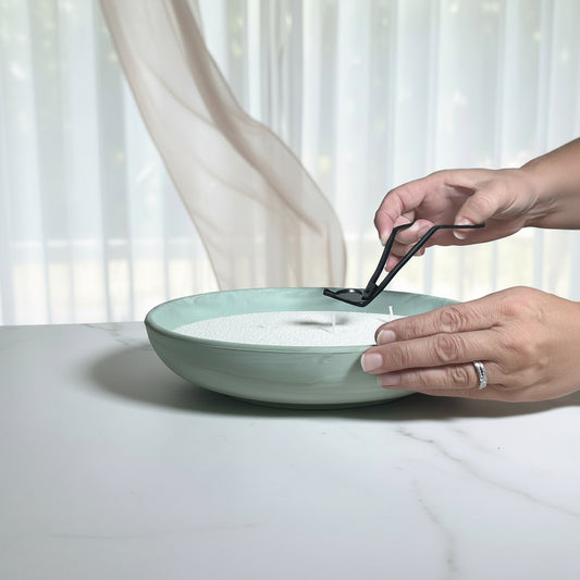 A person's hands are shown lighting a white candle in a shallow, wide ceramic bowl with a sage green rim. The bowl sits on a light wooden table in front of a soft-focus background of white sheer curtains.