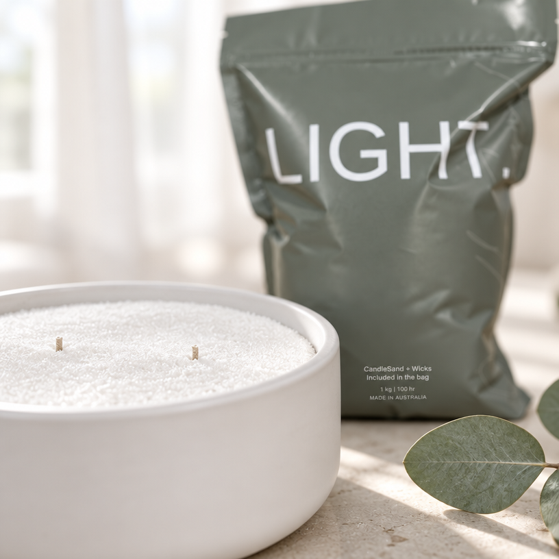 A white ceramic bowl filled with white candlesand wax and two wicks sits in the foreground, with a matte sage green pouch labeled 'LIGHT.' standing behind it on a light surface.