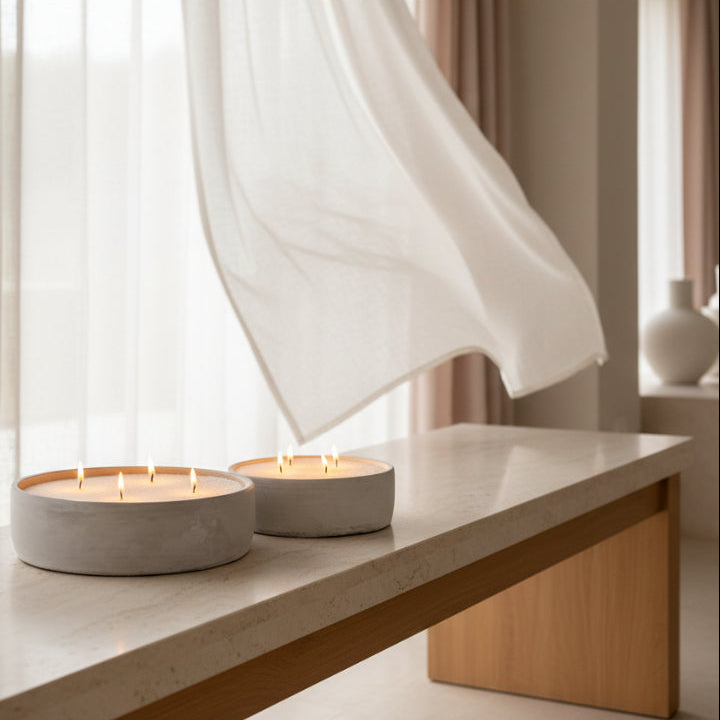 Two round concrete candle holders with lit candles sit on a light stone table in a softly lit interior with sheer white curtains and a wooden base.