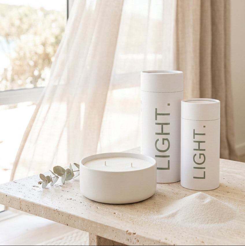 A white ceramic two-wick candle in a round shallow dish sits on a wooden table next to two white cylindrical containers printed with the word 'LIGHT.' in dark green text, against a plain light gray wall.